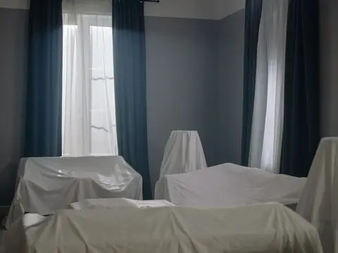 Sofas and chairs under white dust sheets in a room with long curtains